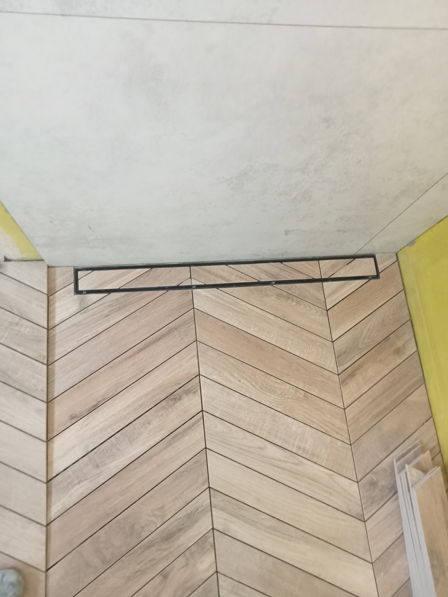 Ukończona podłoga łazienki z jasnych płytek ułożonych w jodełkę, z czarnym odpływem liniowym, widok z góry, fragment ściany z żółtą taśmą malarską.