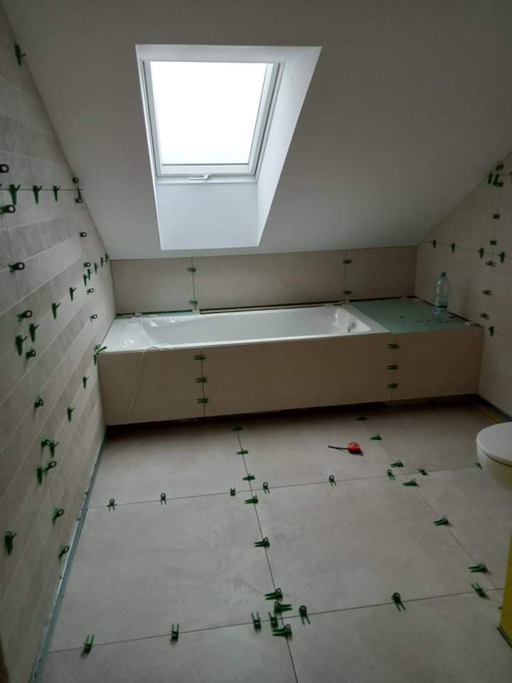 Łazienka na poddaszu w trakcie układania płytek; widoczne krzyżyki dystansowe, wanna zabezpieczona folią, okno dachowe i fragment toalety.