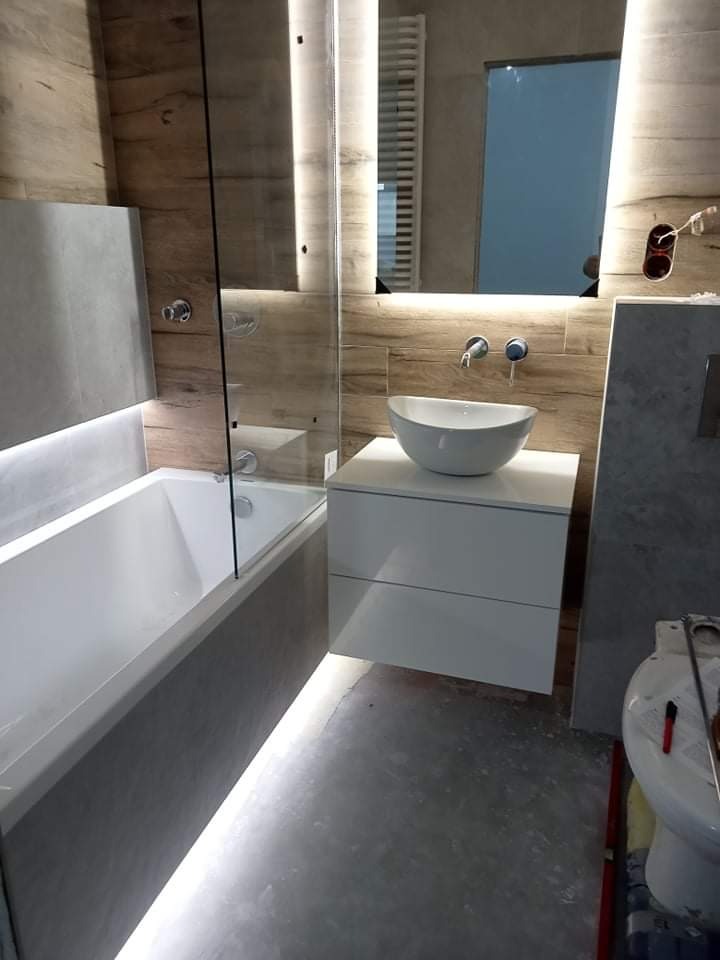 Nowoczesna łazienka z podświetlaną wanną, umywalką nablatową i lustrem z oświetleniem LED, wykończona płytkami imitującymi drewno, w trakcie prac wykończeniowych.