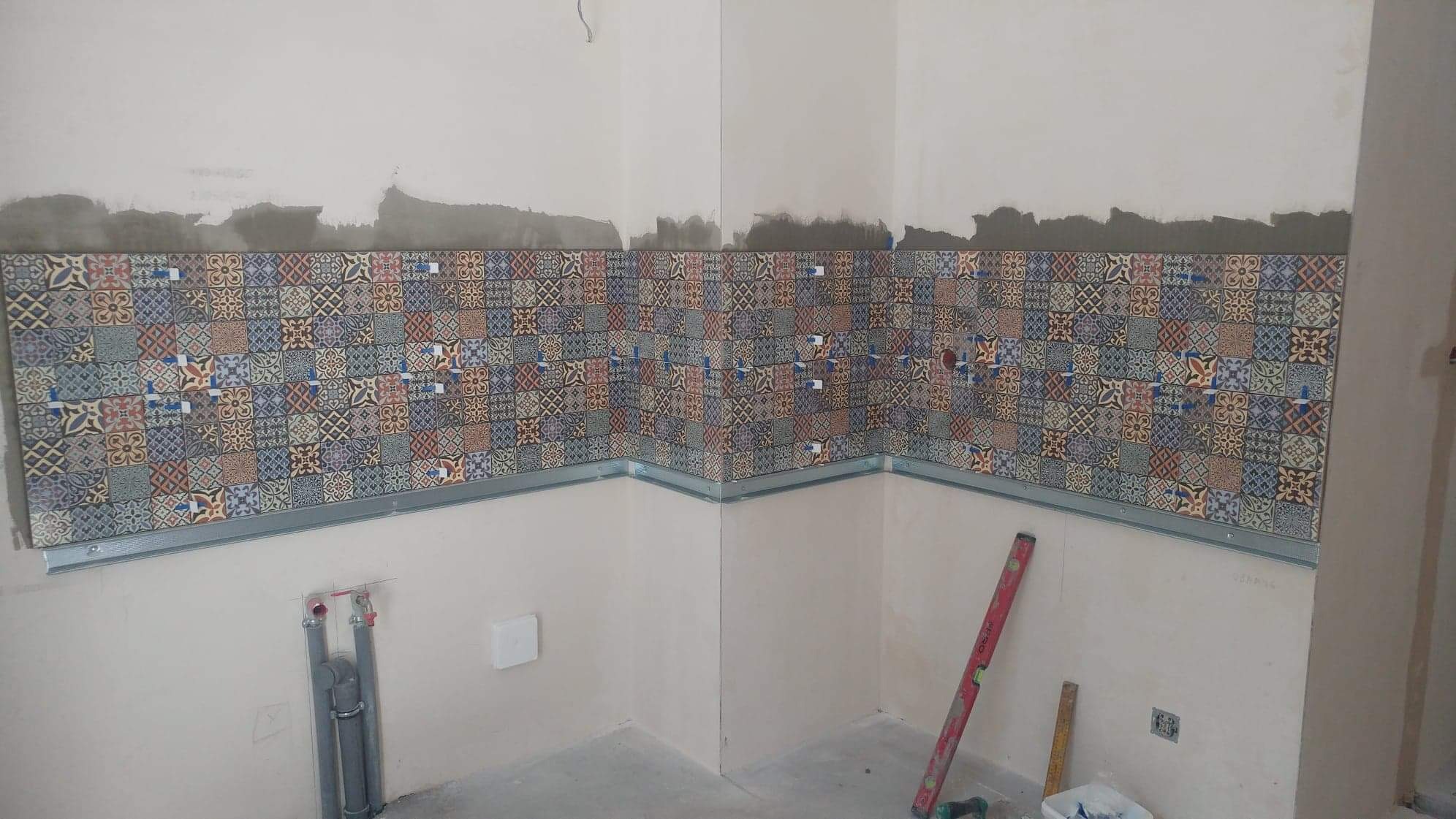 Montaż dekoracyjnych płytek ceramicznych w narożniku pomieszczenia, widoczne profile mocujące i poziomica, ściany częściowo przygotowane do dalszych prac wykończeniowych.