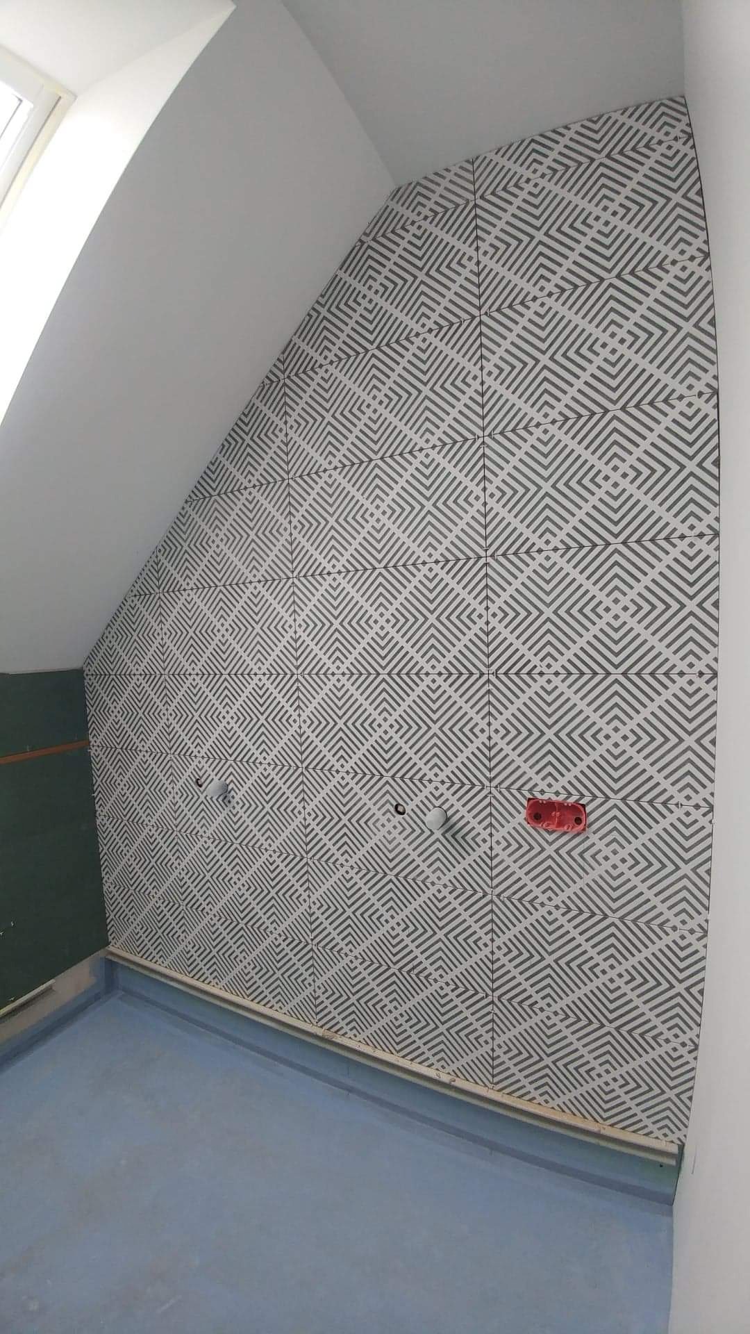 Ściana w łazience na poddaszu wyłożona szaro-białymi płytkami z geometrycznym wzorem, widoczne przygotowane punkty instalacyjne i zabezpieczona podłoga.