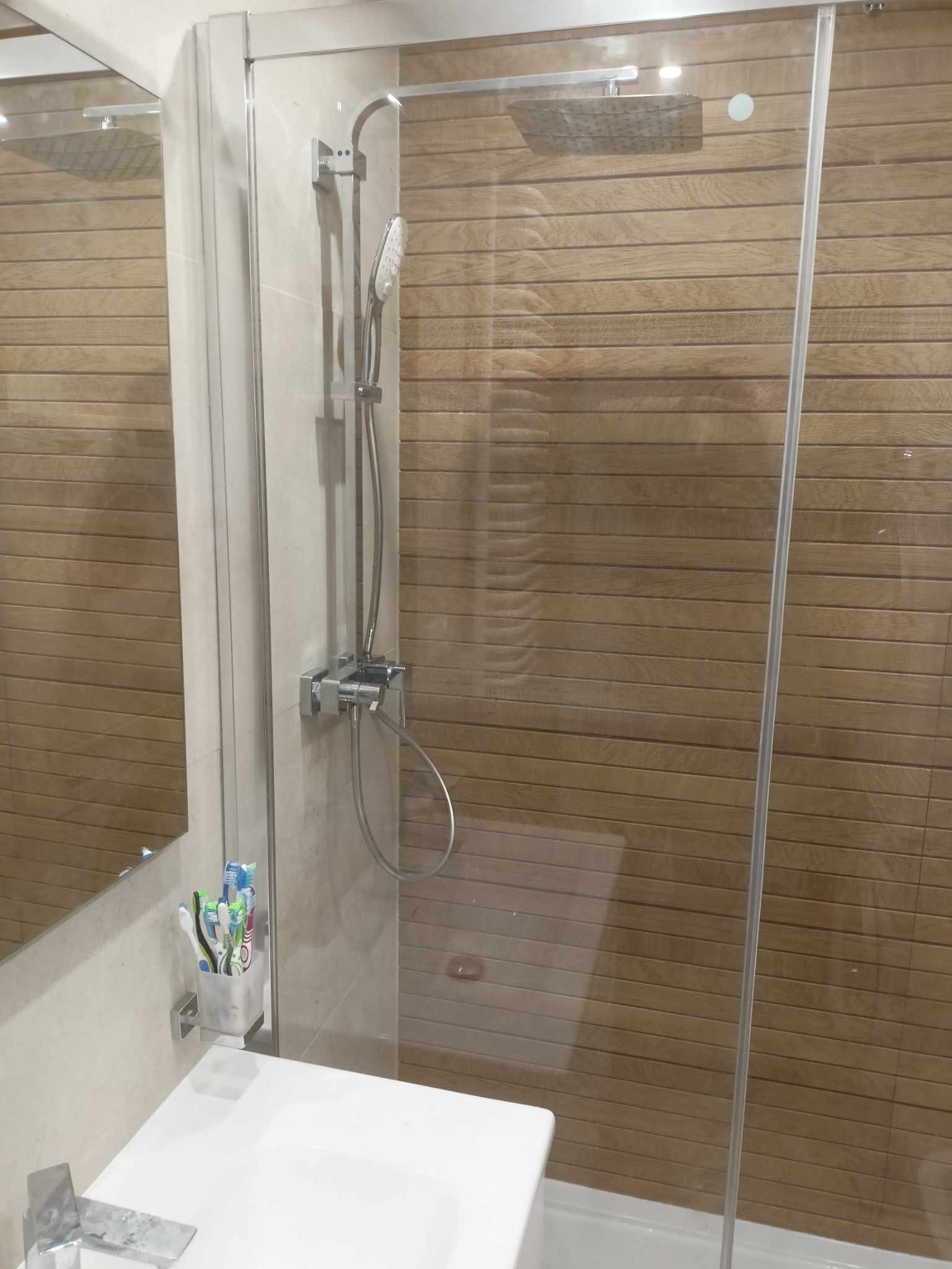 Nowoczesna kabina prysznicowa z deszczownicą i panelami ściennymi imitującymi drewno, widoczne umywalka z baterią i pojemnik na szczoteczki.