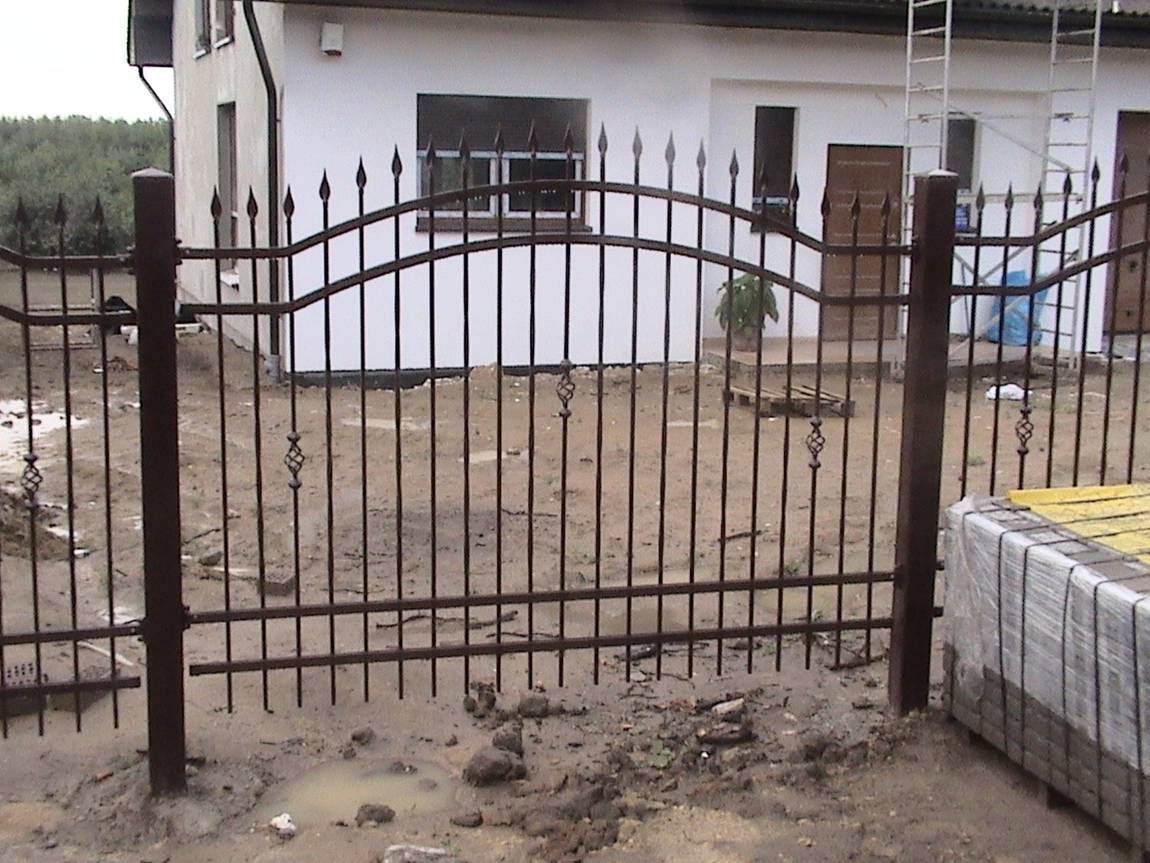 Ciemnobrązowe ogrodzenie metalowe z dekoracyjnymi elementami w kształcie koszyczków, zakończone grotami, na tle białego domu w trakcie budowy, widoczne kałuże i materiały budowlane.