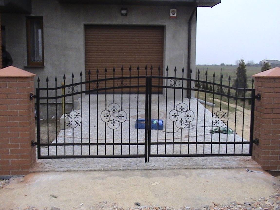 Dwuskrzydłowa, czarna, metalowa brama z ozdobnymi elementami kwiatowymi, osadzona między murowanymi słupkami z cegły klinkierowej, na tle szarego budynku z brązową bramą garażową.