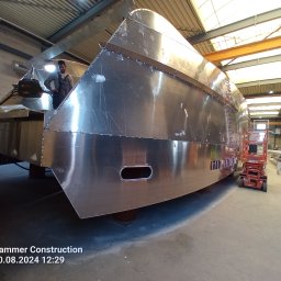 Firma Usługowa Marcin Jażdżewski - Metalowa konstrukcja łodzi w hali produkcyjnej, pracownik w kombinezonie nadzoruje proces, widoczny podnośnik i elementy konstrukcyjne hali.