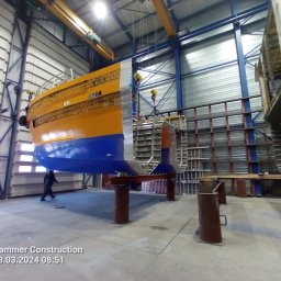 Firma Usługowa Marcin Jażdżewski - Fragment konstrukcji statku w hali produkcyjnej, podwieszony na suwnicy, widoczny pracownik w tle.