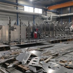 Firma Usługowa Marcin Jażdżewski - Metalowe konstrukcje szkieletowe z otworami, ustawione w hali produkcyjnej, z widocznymi odpadami metalowymi na pierwszym planie i pracownikami w tle.