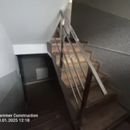 Klatka schodowa z drewnianymi stopniami i prostą, metalową balustradą, widok z góry na dół schodów.