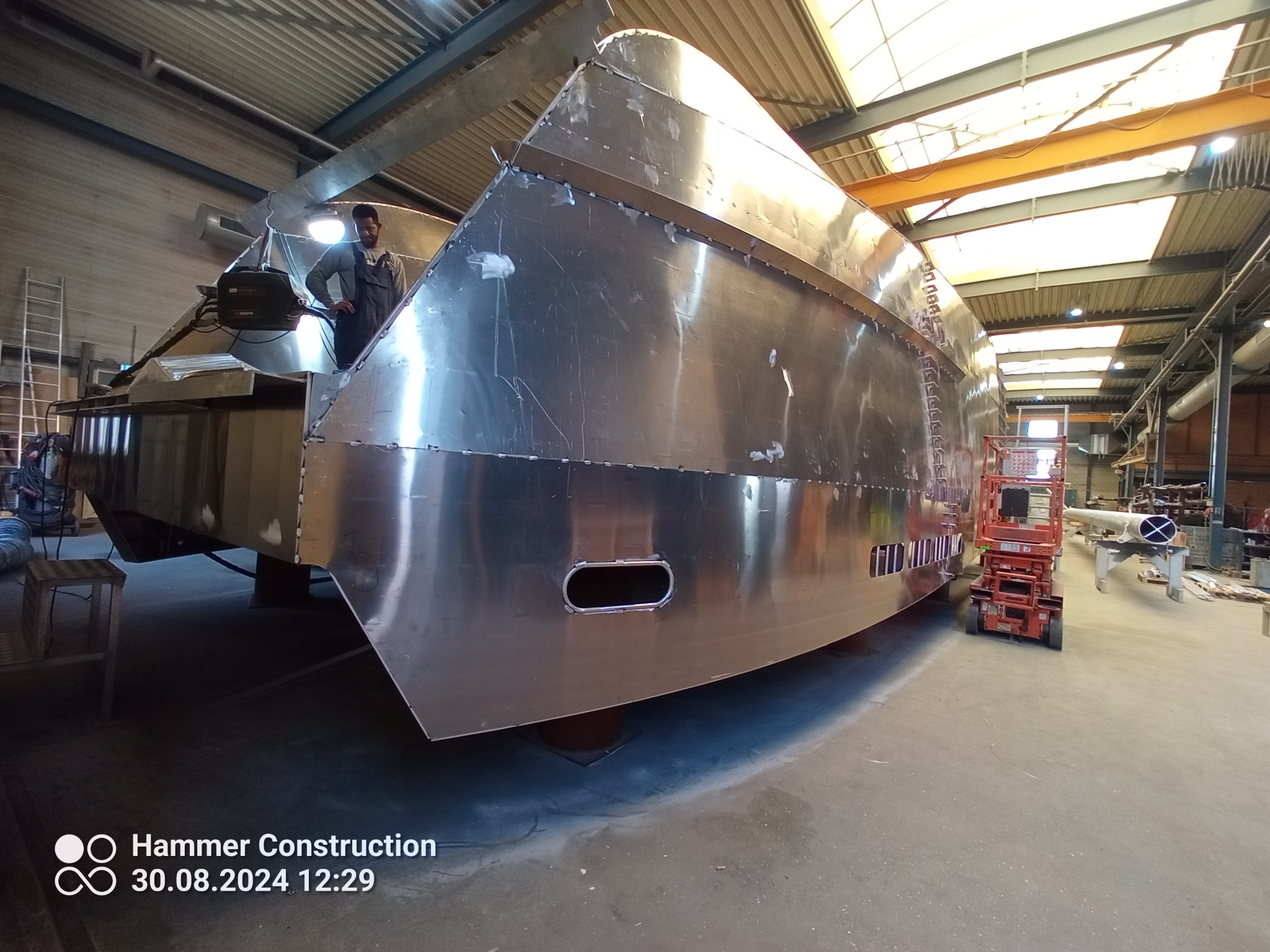 Metalowa konstrukcja łodzi w hali produkcyjnej, pracownik w kombinezonie nadzoruje proces, widoczny podnośnik i elementy konstrukcyjne hali.