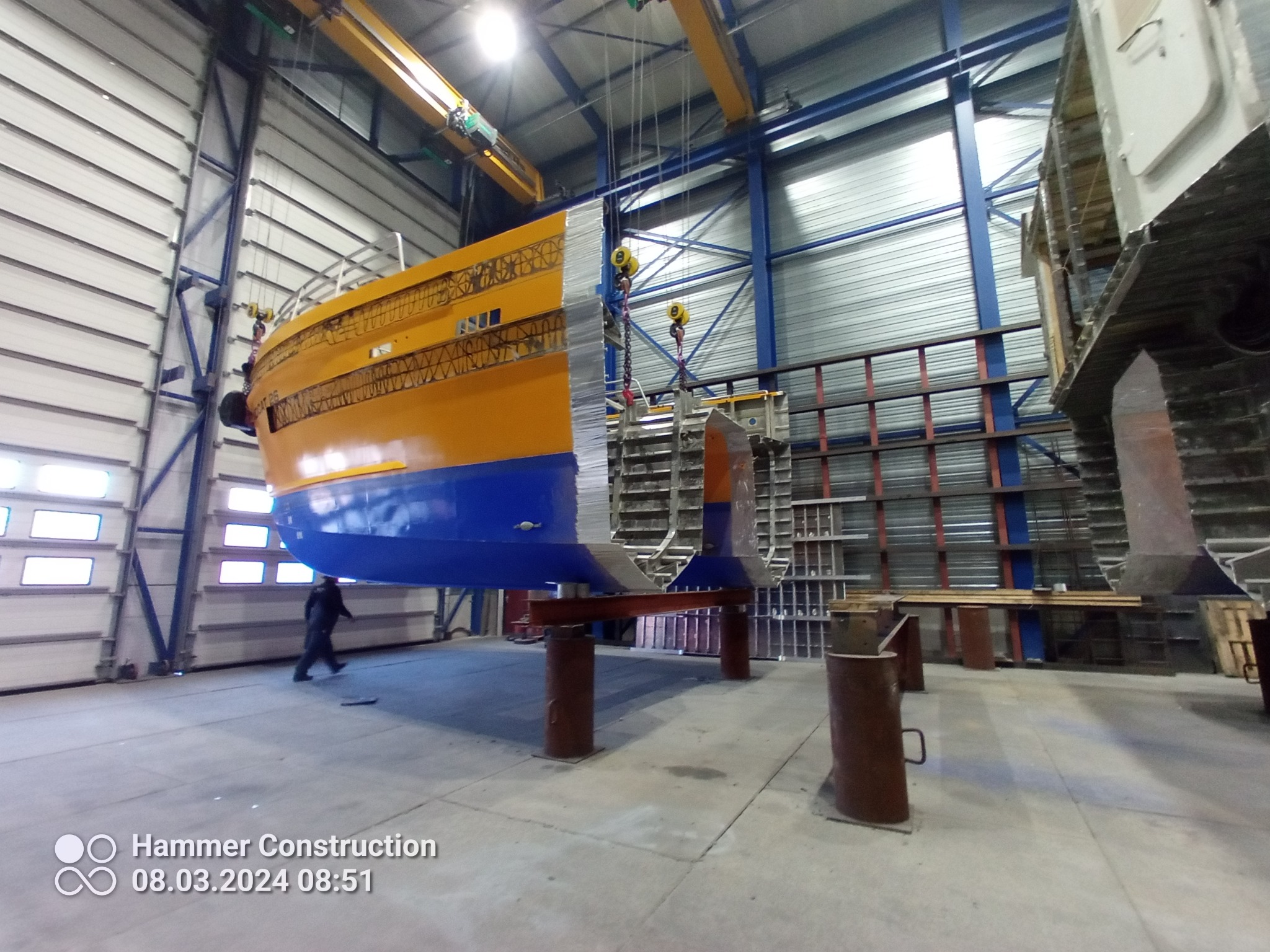 Fragment konstrukcji statku w hali produkcyjnej, podwieszony na suwnicy, widoczny pracownik w tle.