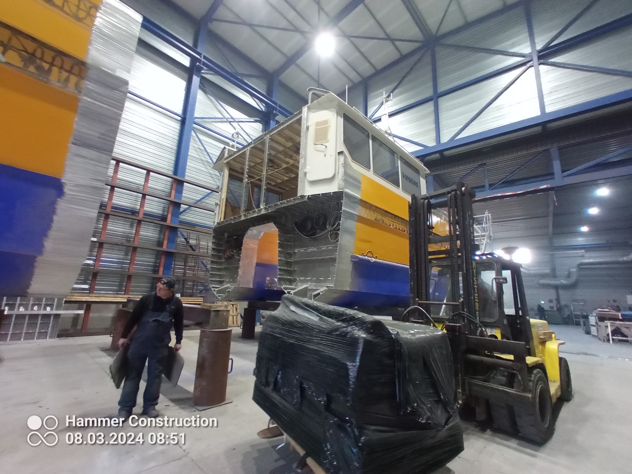 Konstrukcja stalowa modułu budynku w hali produkcyjnej, transportowana wózkiem widłowym, pracownik w ubraniu roboczym w tle.