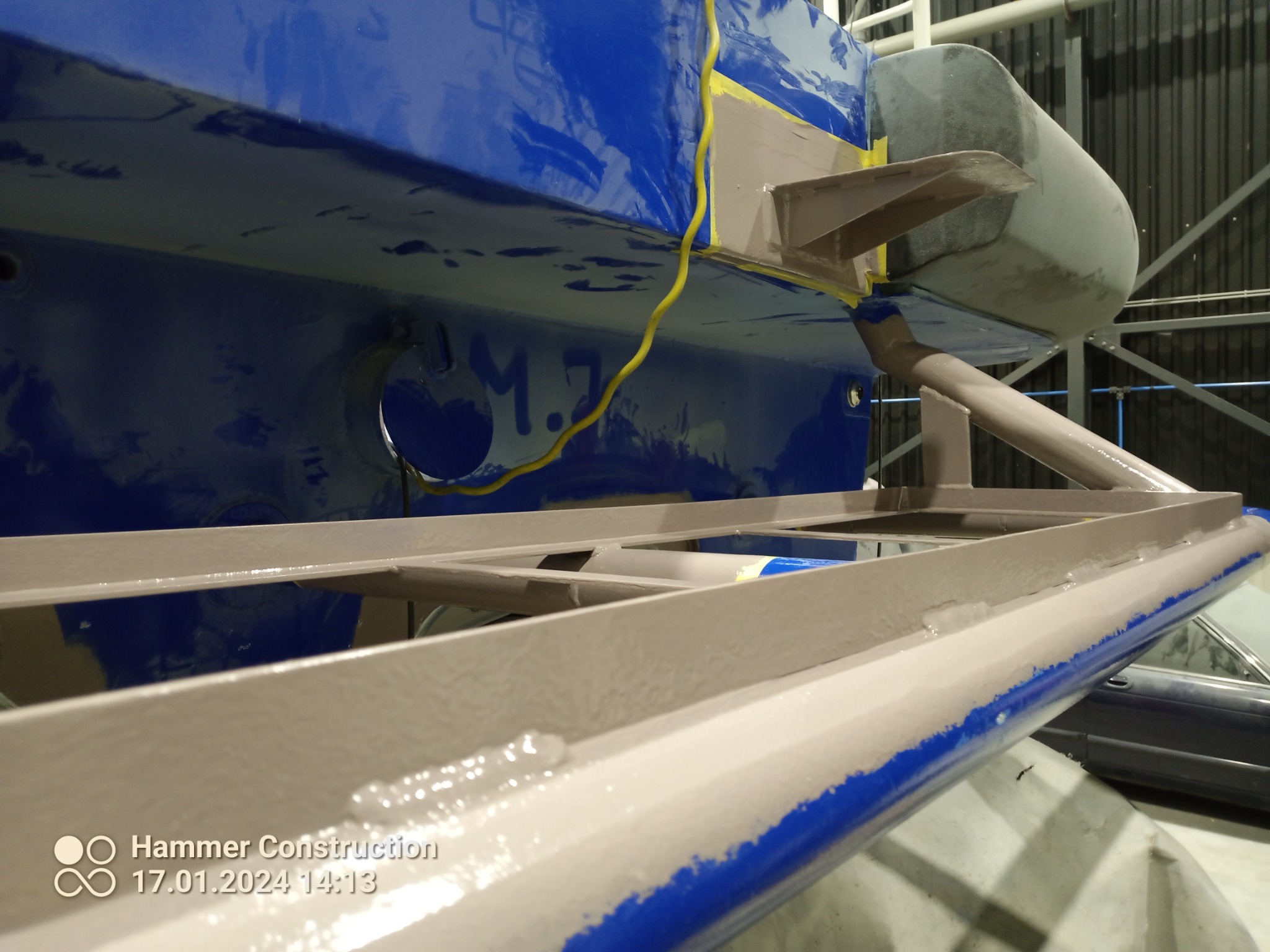 Fragment konstrukcji metalowej z elementami pomalowanymi na szaro i niebiesko, w trakcie prac wykończeniowych, widoczny fragment żółtego kabla i oznaczenie 'M.7'.