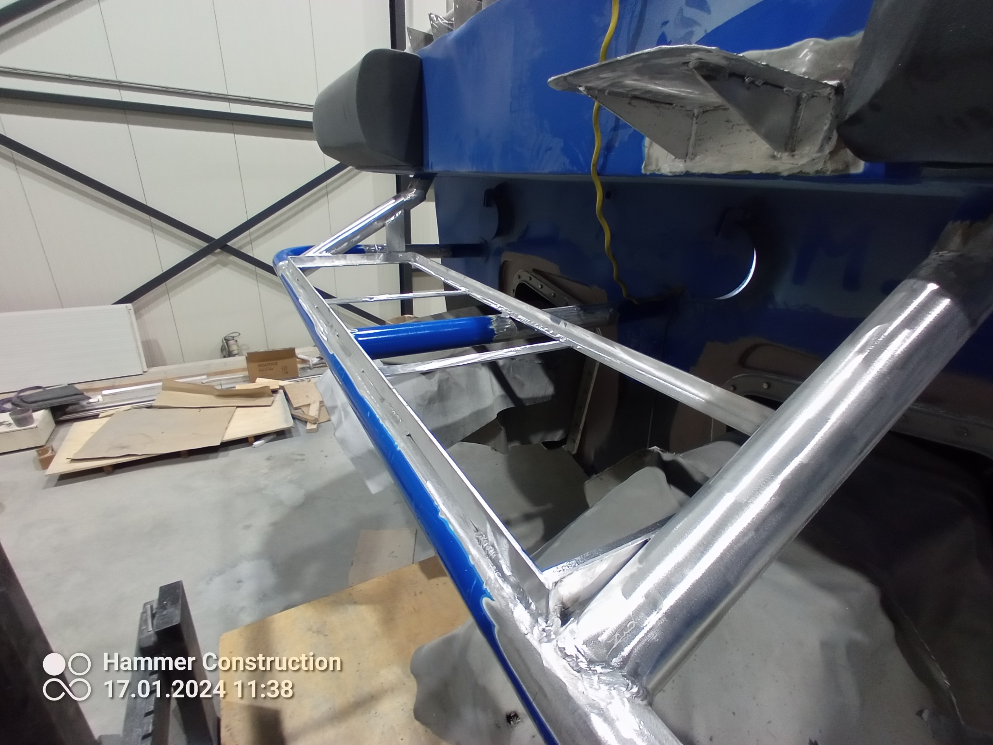 Spawana konstrukcja z rur, prawdopodobnie element ramy lub podwozia, z widocznymi spawami i niebieskim akcentem kolorystycznym, w hali produkcyjnej.