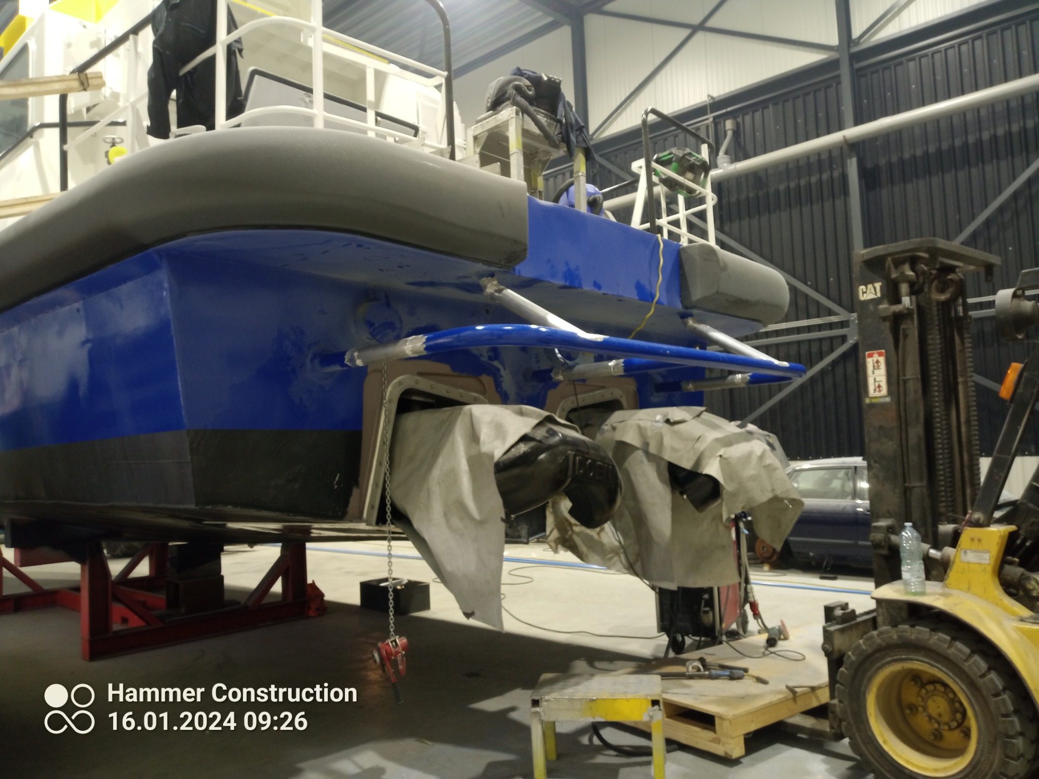 Dolna część niebieskiej łodzi na stojakach w hali, z widocznymi elementami konstrukcyjnymi i osłoniętymi śrubami, obok wózek widłowy i narzędzia.