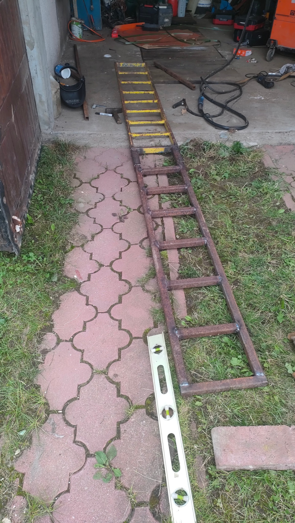 Rampa z żółtymi szczeblami i surowa metalowa konstrukcja drabiny leżą na kostce brukowej obok poziomicy; widoczne ślady spawania i narzędzia w tle.