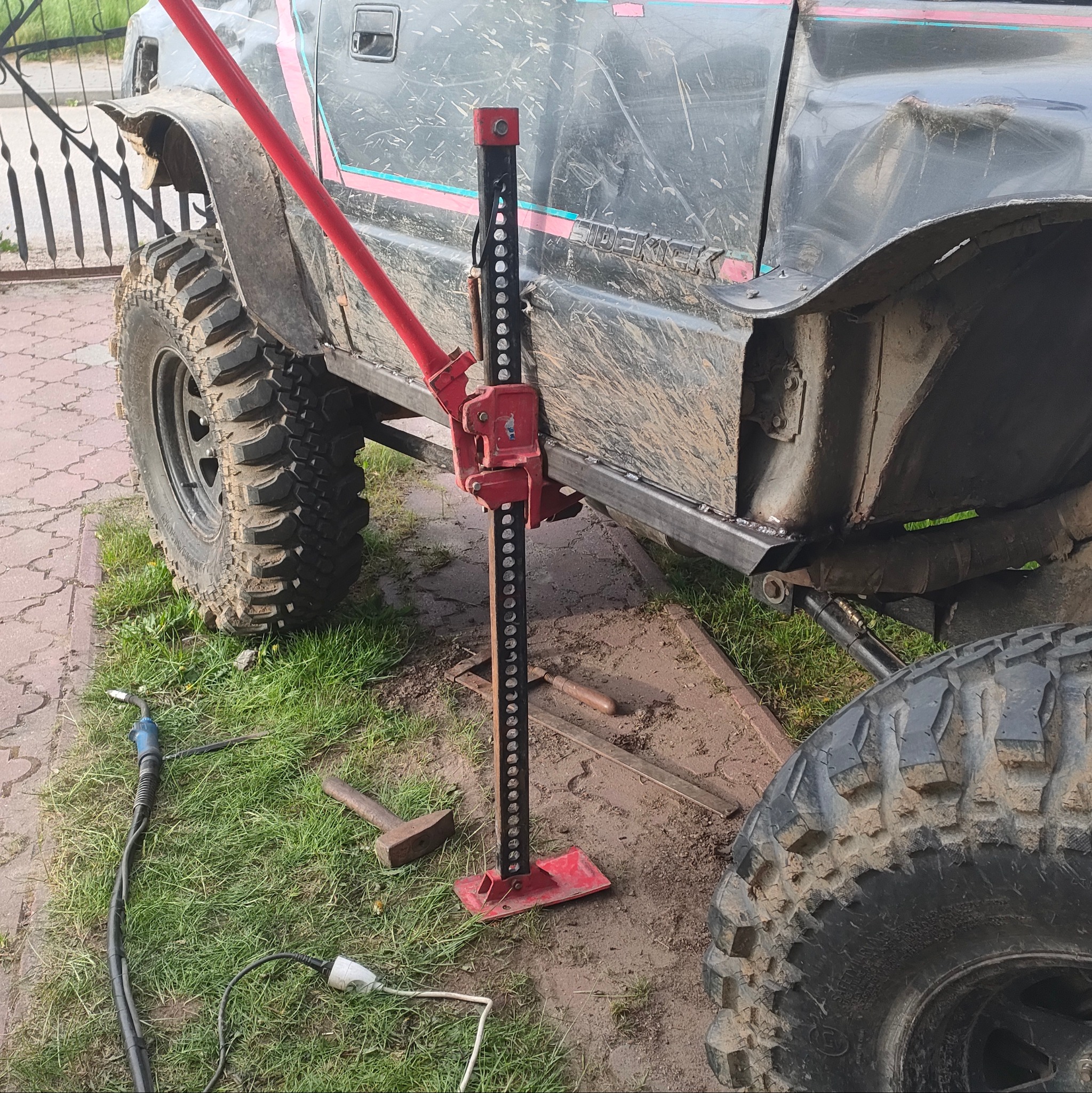 Częściowo naprawione podwozie brudnego samochodu terenowego podniesione czerwonym podnośnikiem typu hi-lift, z widocznym spawem mocującym element wzmacniający