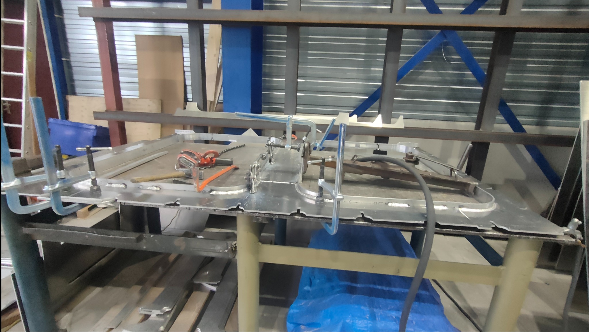 Metalowa konstrukcja z elementami mocującymi, narzędzia i spawarka na stole warsztatowym w hali produkcyjnej.