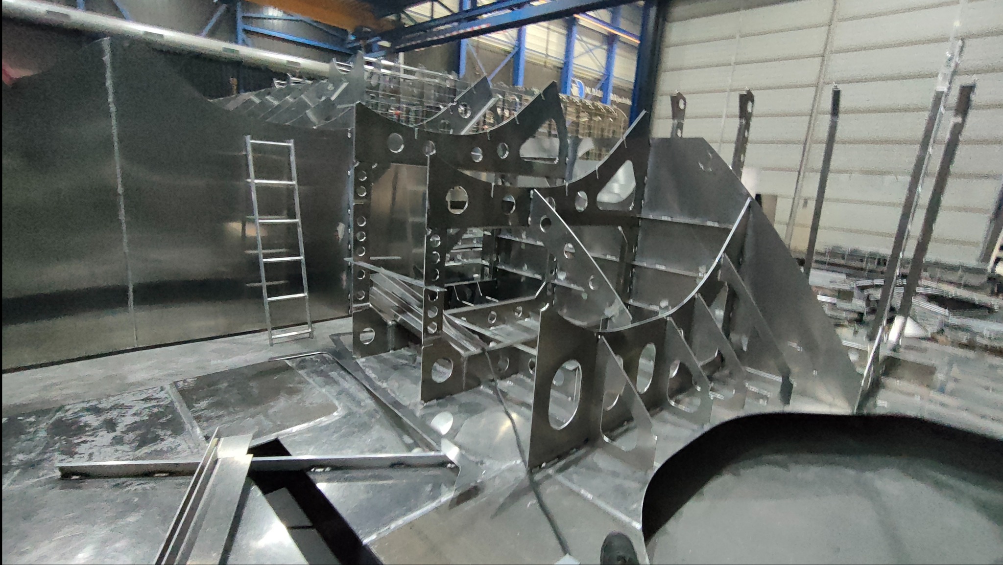 Metalowa konstrukcja szkieletowa z licznymi otworami, przypominająca kadłub statku, w hali produkcyjnej z metalową ścianą i drabiną w tle.