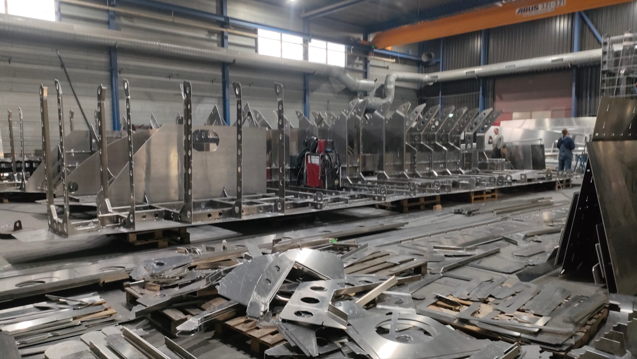 Metalowe konstrukcje szkieletowe z otworami, ustawione w hali produkcyjnej, z widocznymi odpadami metalowymi na pierwszym planie i pracownikami w tle.