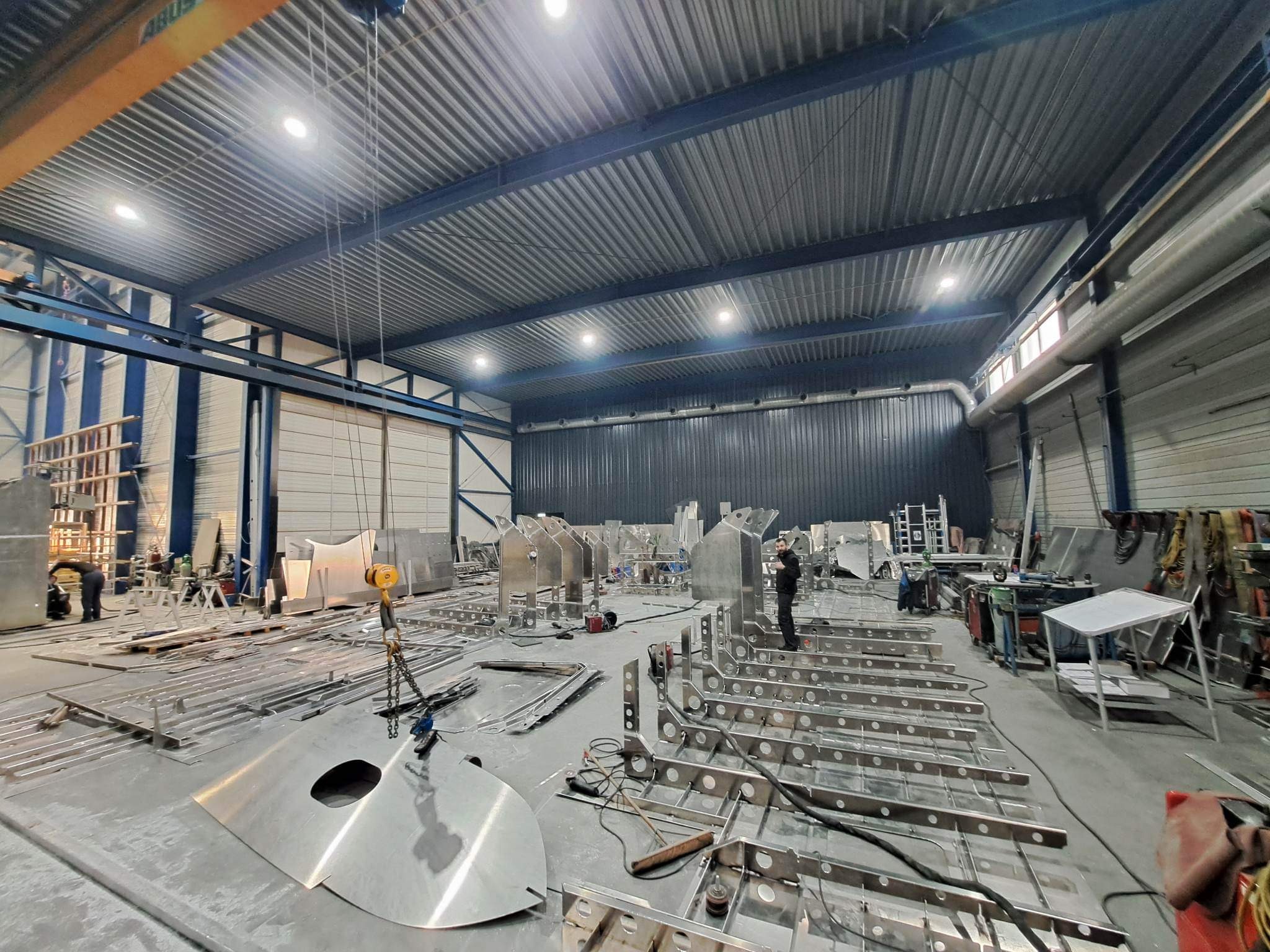 Warsztat z surowymi elementami konstrukcji ze stali nierdzewnej, widoczny suwnica i pracownik w oddali, przygotowanie do spawania lub montażu.