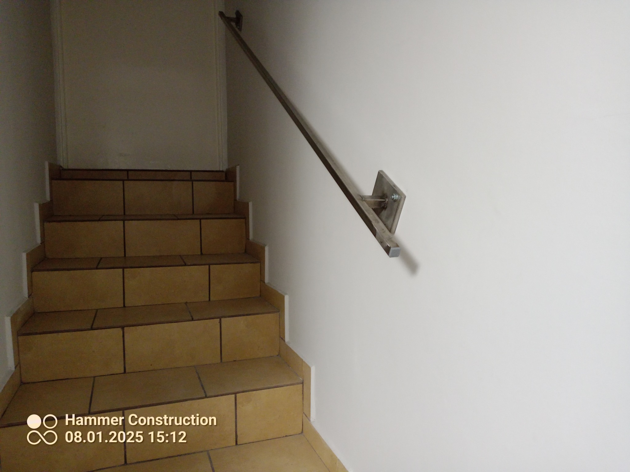 Metalowa poręcz schodowa przymocowana do ściany, widoczne mocowania na śrubach, brązowe płytki na schodach.