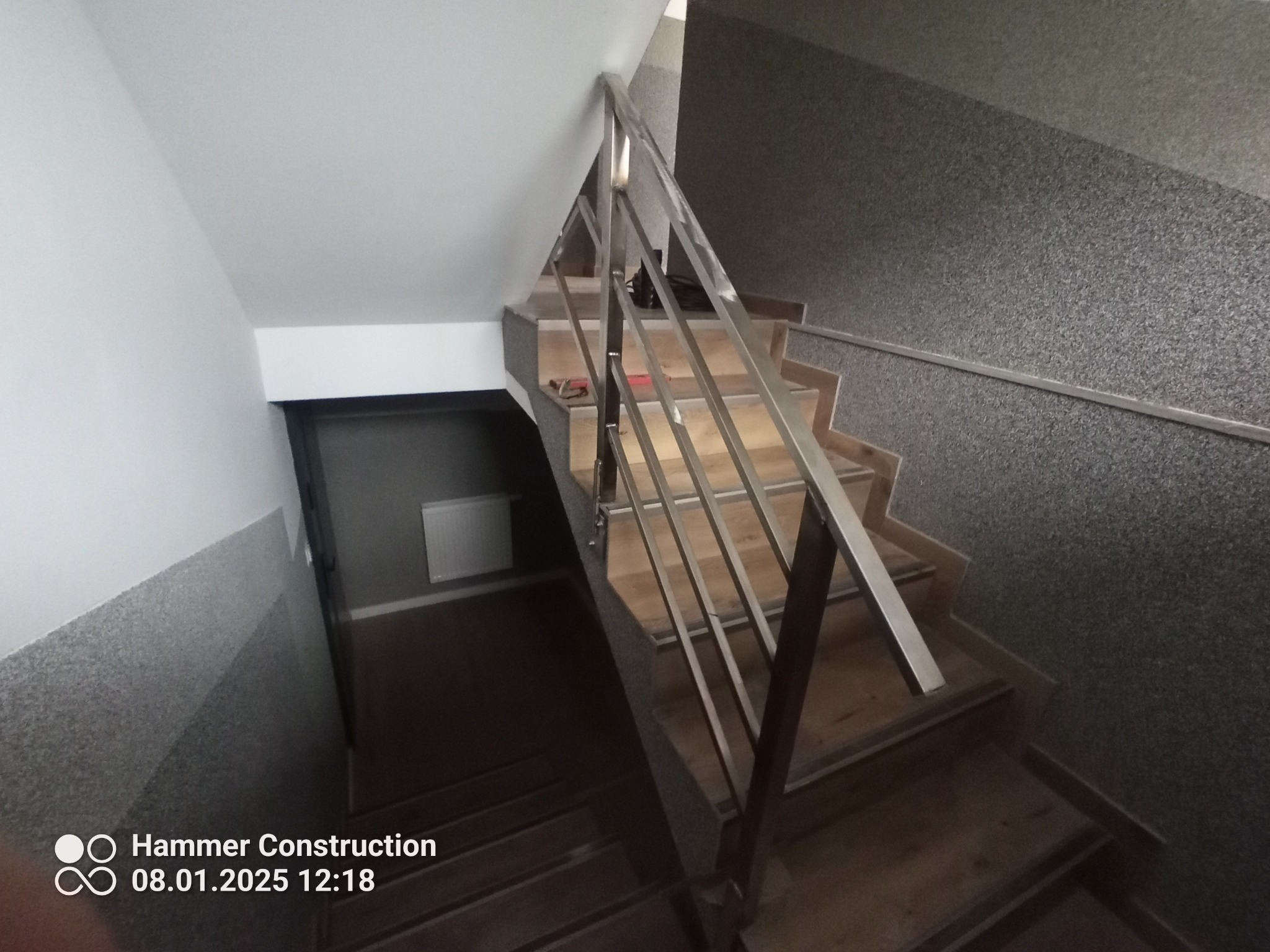 Klatka schodowa z drewnianymi stopniami i prostą, metalową balustradą, widok z góry na dół schodów.