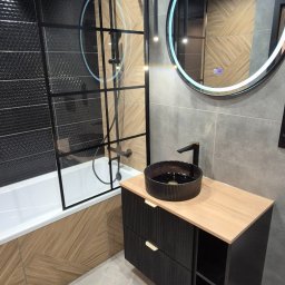 Konrad Szlaga - Nowoczesna łazienka z czarną armaturą, kabiną prysznicową typu walk-in i okrągłym lustrem z podświetleniem LED. Umywalka nablatowa na szafce z drewnianym blatem.
