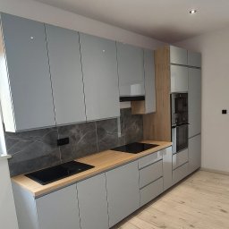 Konrad Szlaga - Nowoczesna kuchnia z szarymi, lakierowanymi frontami, drewnianym blatem i marmurowym backsplashiem. Widoczne płyty indukcyjne i zabudowany piekarnik.