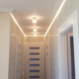 Konrad Szlaga - Korytarz po remoncie: białe ściany z efektem dekoracyjnym, podświetlany sufit z listwami i trzema lampami, białe drzwi z niebieskimi akcentami. Widok z dołu.