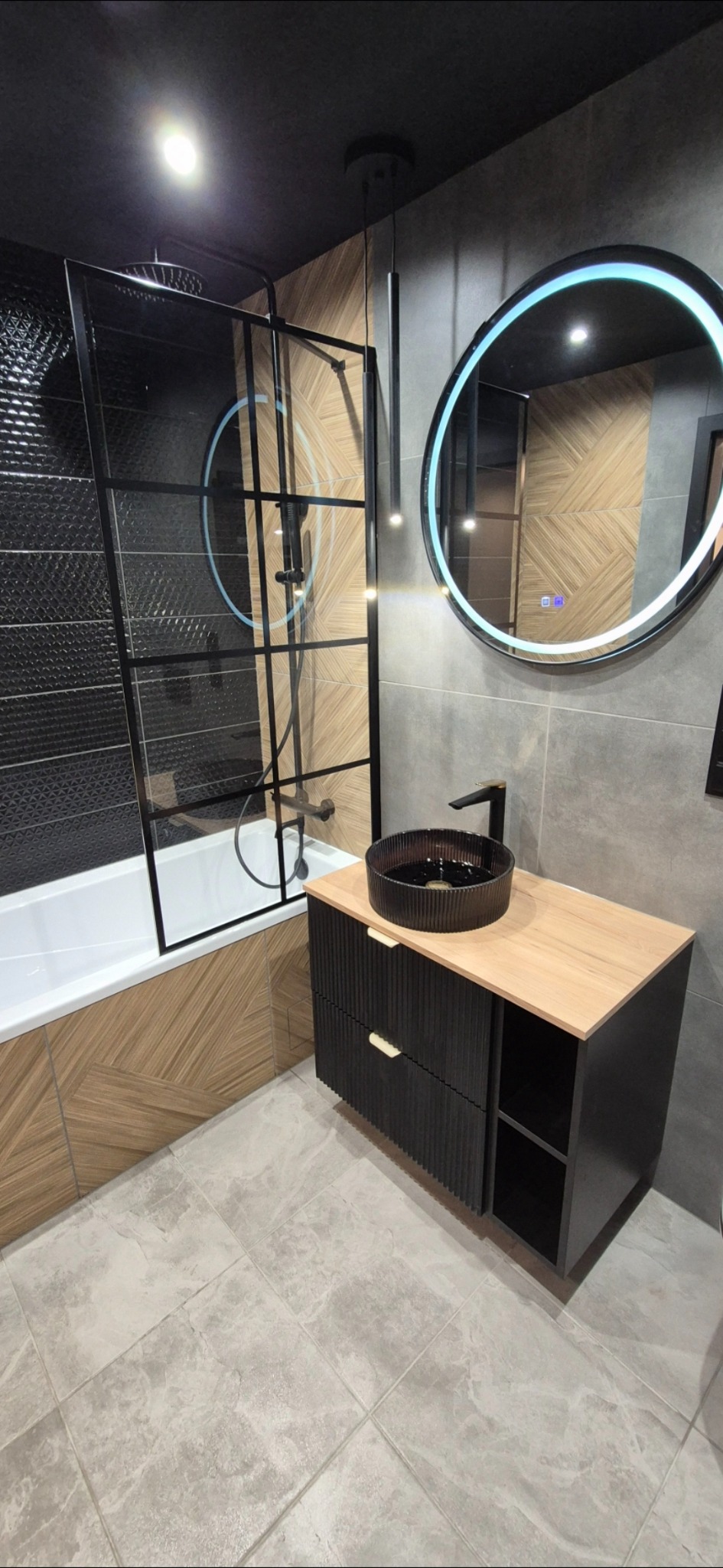 Nowoczesna łazienka z czarną armaturą, kabiną prysznicową typu walk-in i okrągłym lustrem z podświetleniem LED. Umywalka nablatowa na szafce z drewnianym blatem.