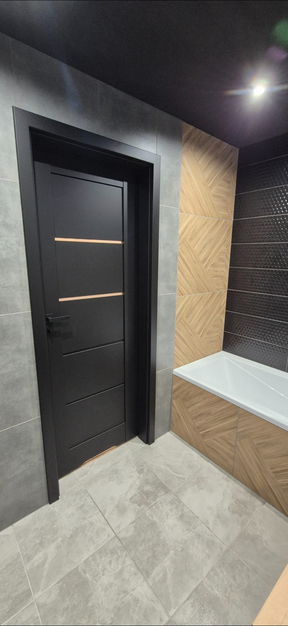 Ciemna, nowoczesna łazienka z czarnymi drzwiami z poziomymi wstawkami, szarą płytką ścienną i podłogową oraz dekoracyjną okładziną w strefie wanny.
