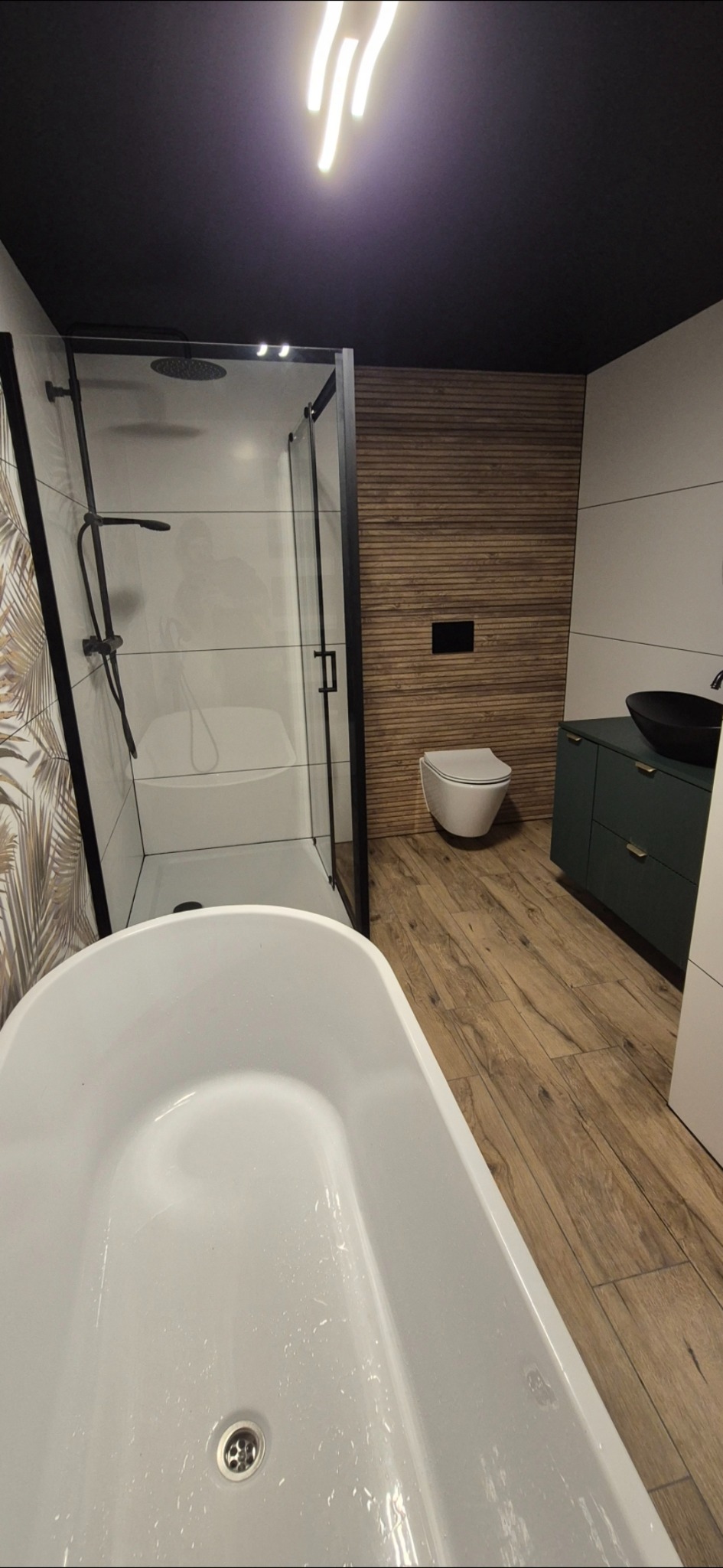Nowoczesna łazienka z wanną wolnostojącą, kabiną prysznicową typu walk-in, drewnianą okładziną ściany i podłogą imitującą drewno. Czarny sufit i armatura.