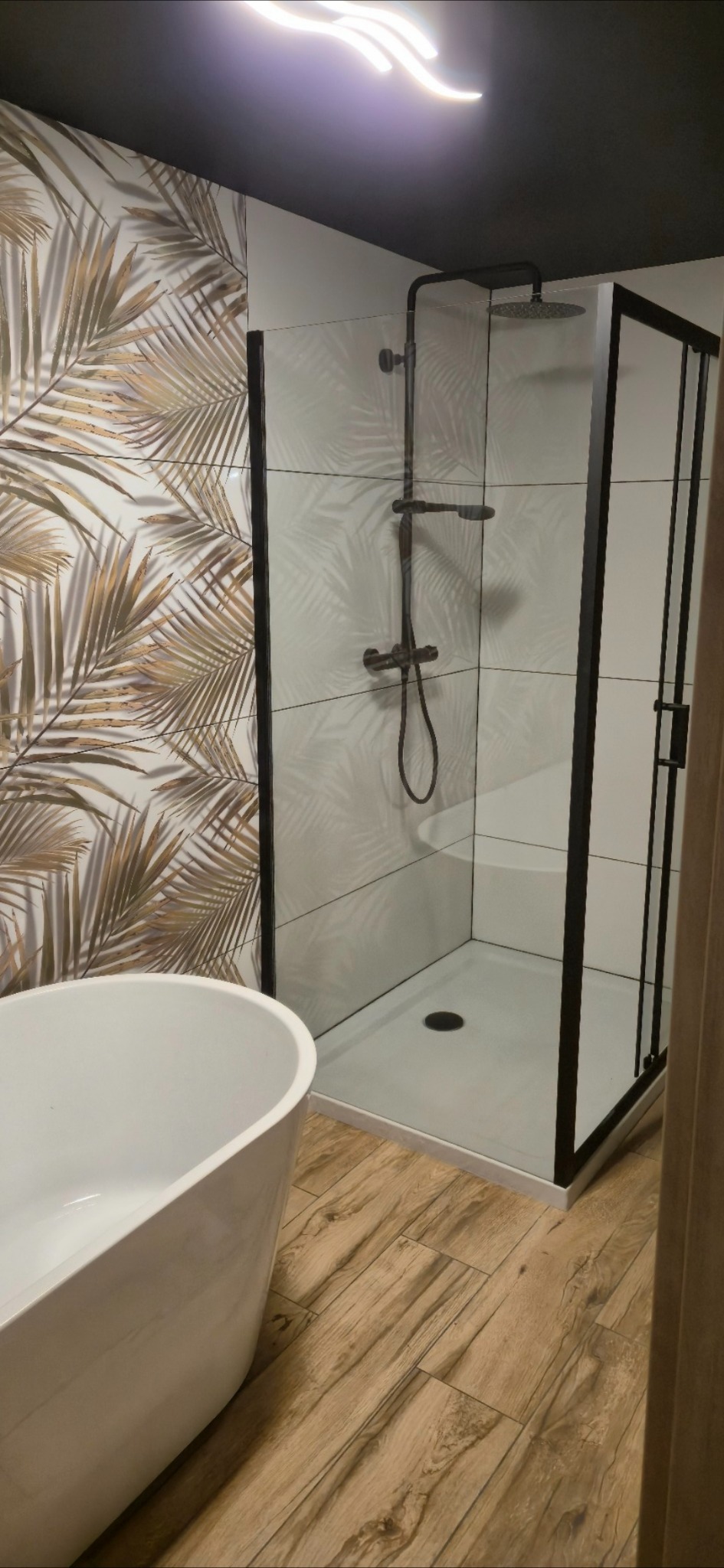 Nowoczesna łazienka z kabiną prysznicową typu walk-in z czarnymi ramami, wolnostojącą wanną i płytkami z motywem liści palmowych. Drewnopodobna podłoga.