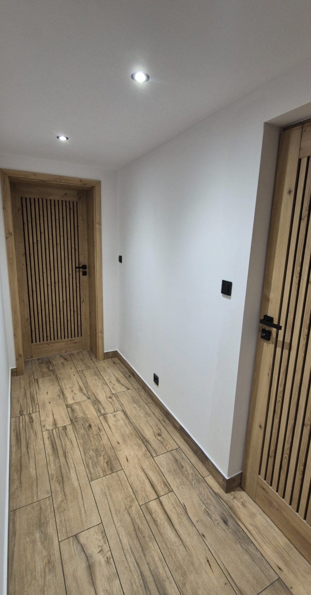 Nowoczesny korytarz z drewnopodobną podłogą, białymi ścianami i dwiema drewnianymi drzwiami z czarnymi klamkami. Oświetlenie punktowe w suficie.