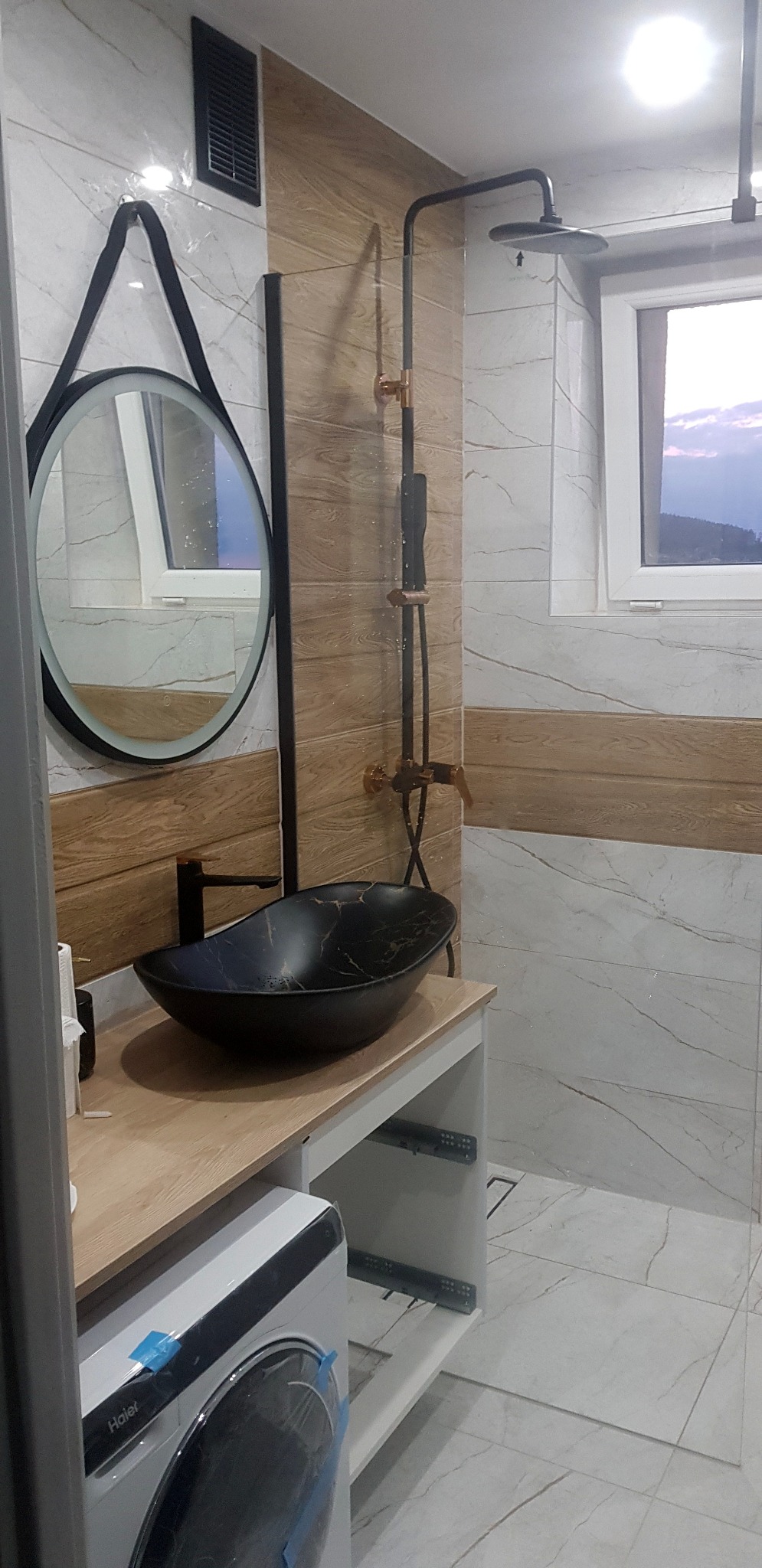 Nowoczesna łazienka z czarną umywalką na blacie z jasnego drewna, okrągłym lustrem w czarnej ramie i kabiną prysznicową typu walk-in z deszczownicą.