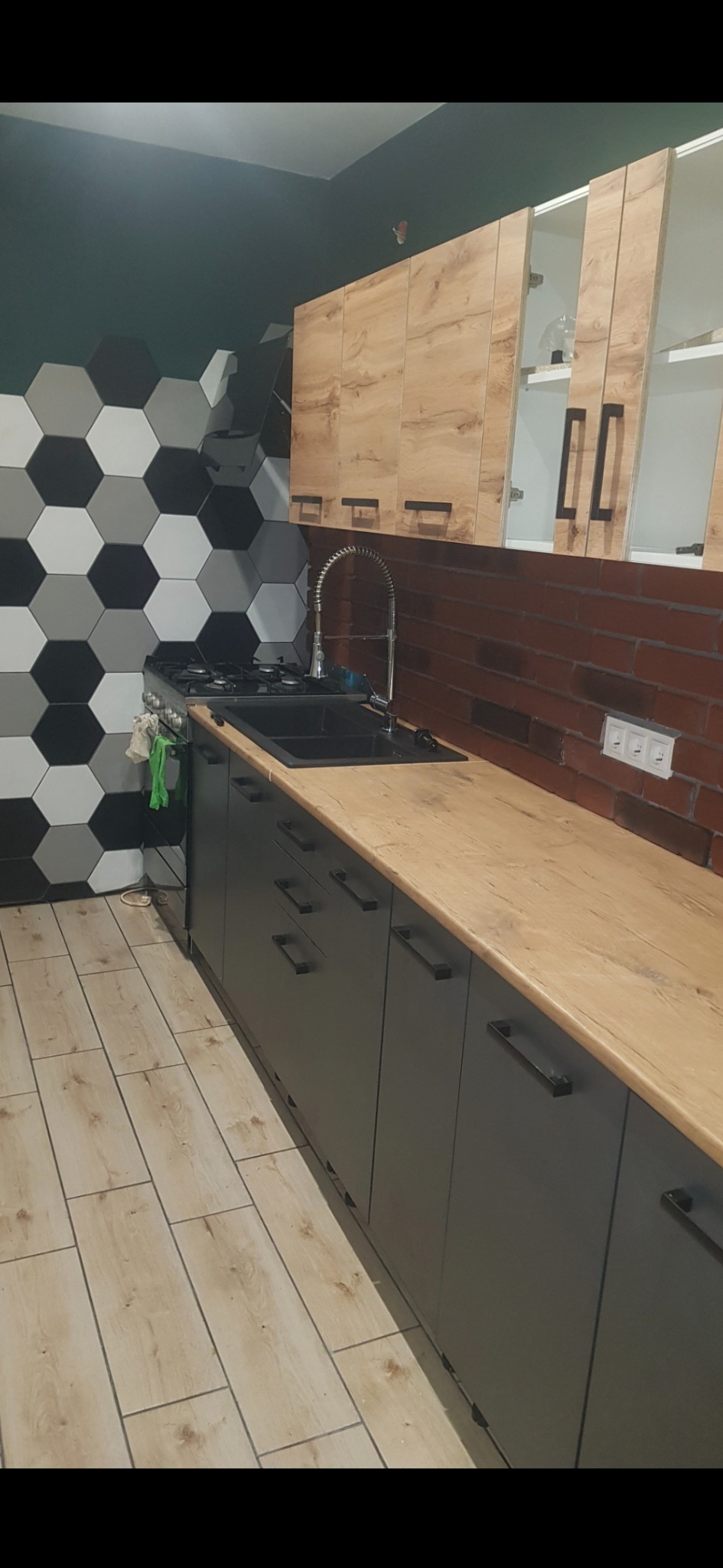 Nowoczesna kuchnia po remoncie: szare szafki z drewnianym blatem, heksagonalne płytki na ścianie i ceglana mozaika nad blatem. Widok na wykończone wnętrze.