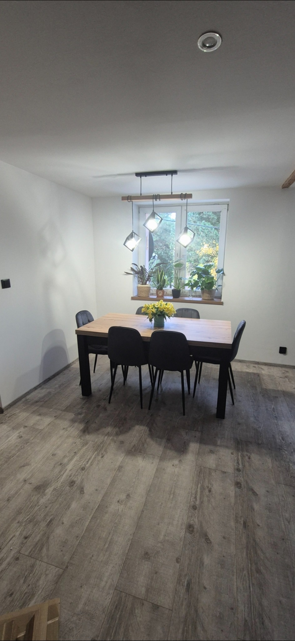 Jasne wnętrze jadalni z drewnianą podłogą, stołem z bukietem żółtych kwiatów i nowoczesną lampą wiszącą. Za oknem widok na zieleń. Minimalistyczny design.