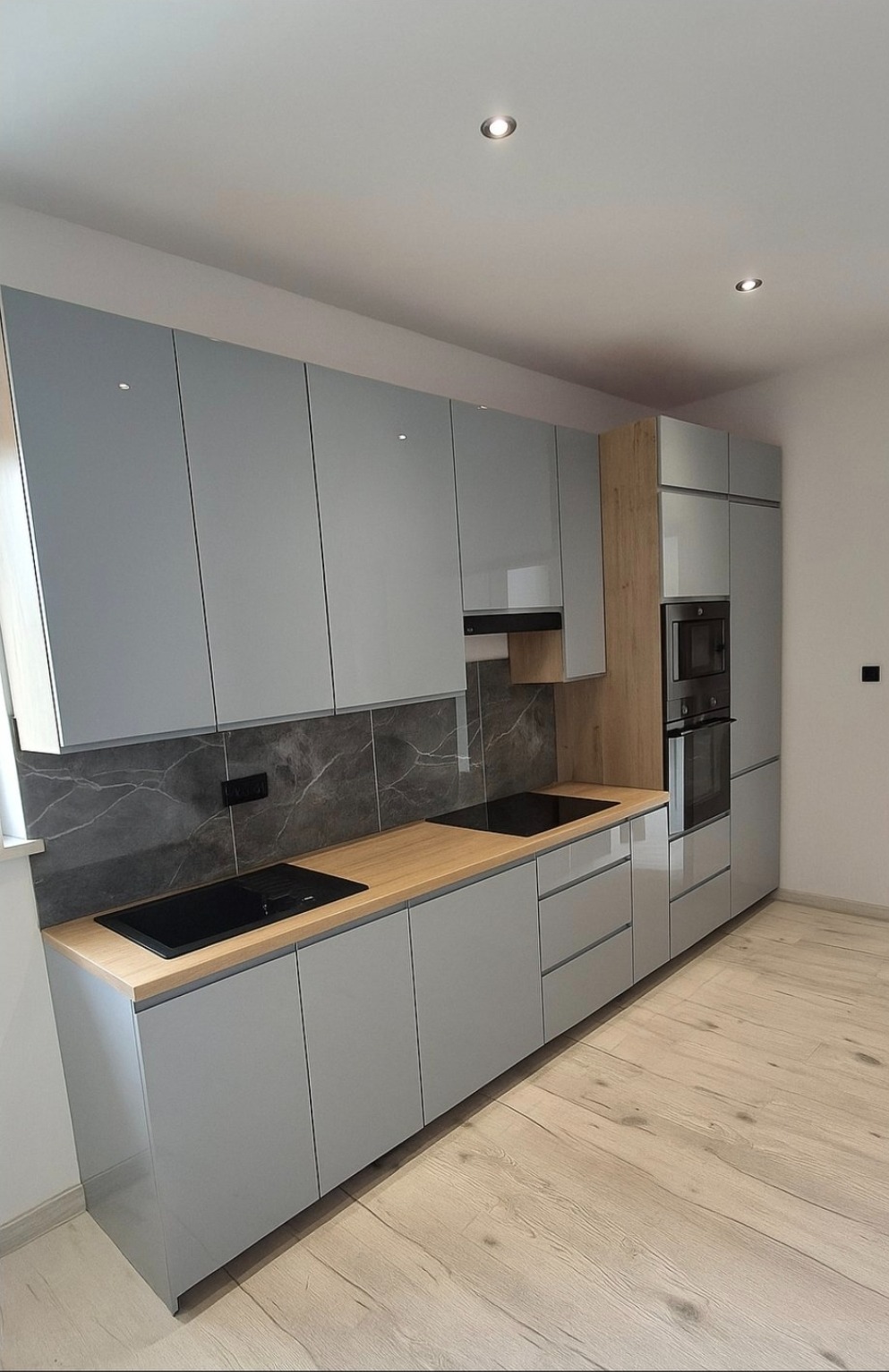 Nowoczesna kuchnia z szarymi, lakierowanymi frontami, drewnianym blatem i marmurowym backsplashiem. Widoczne płyty indukcyjne i zabudowany piekarnik.