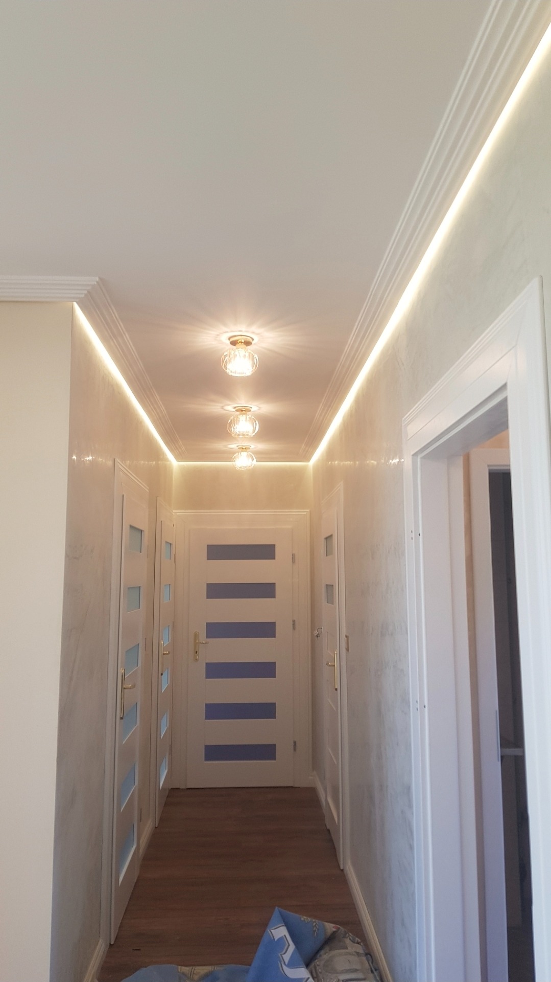 Korytarz po remoncie: białe ściany z efektem dekoracyjnym, podświetlany sufit z listwami i trzema lampami, białe drzwi z niebieskimi akcentami. Widok z dołu.
