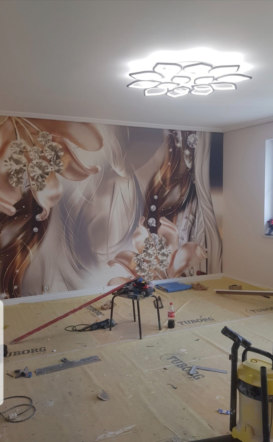 Pokój w trakcie tapetowania z motywem kwiatowym. Widoczne narzędzia, podłoga zabezpieczona folią, nowoczesna lampa sufitowa. Ujęcie z poziomu podłogi.