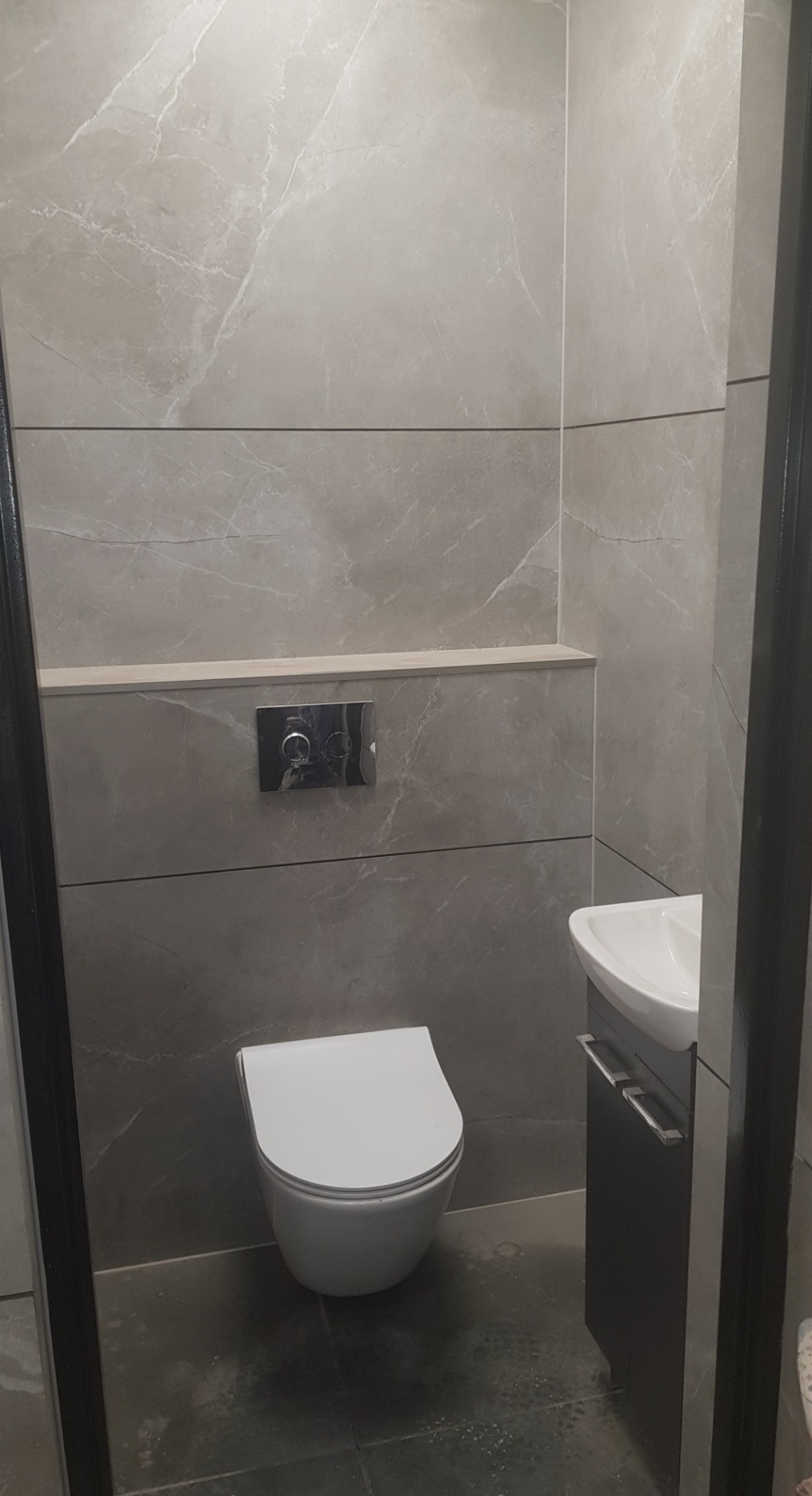 Nowoczesna łazienka z szarymi płytkami imitującymi marmur, podwieszana toaleta i umywalka z szafką. Minimalistyczny design z chromowanymi detalami.