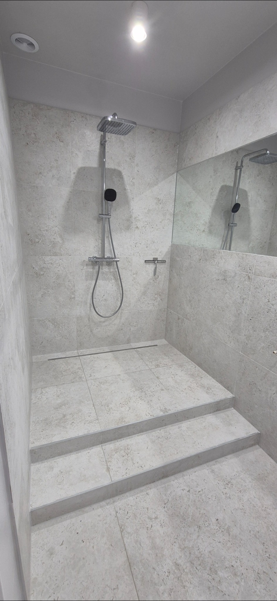 Wykończona kabina prysznicowa z dwoma stopniami, wyłożona jasnymi płytkami imitującymi kamień. Widoczna deszczownica, odpływ liniowy i lustro.