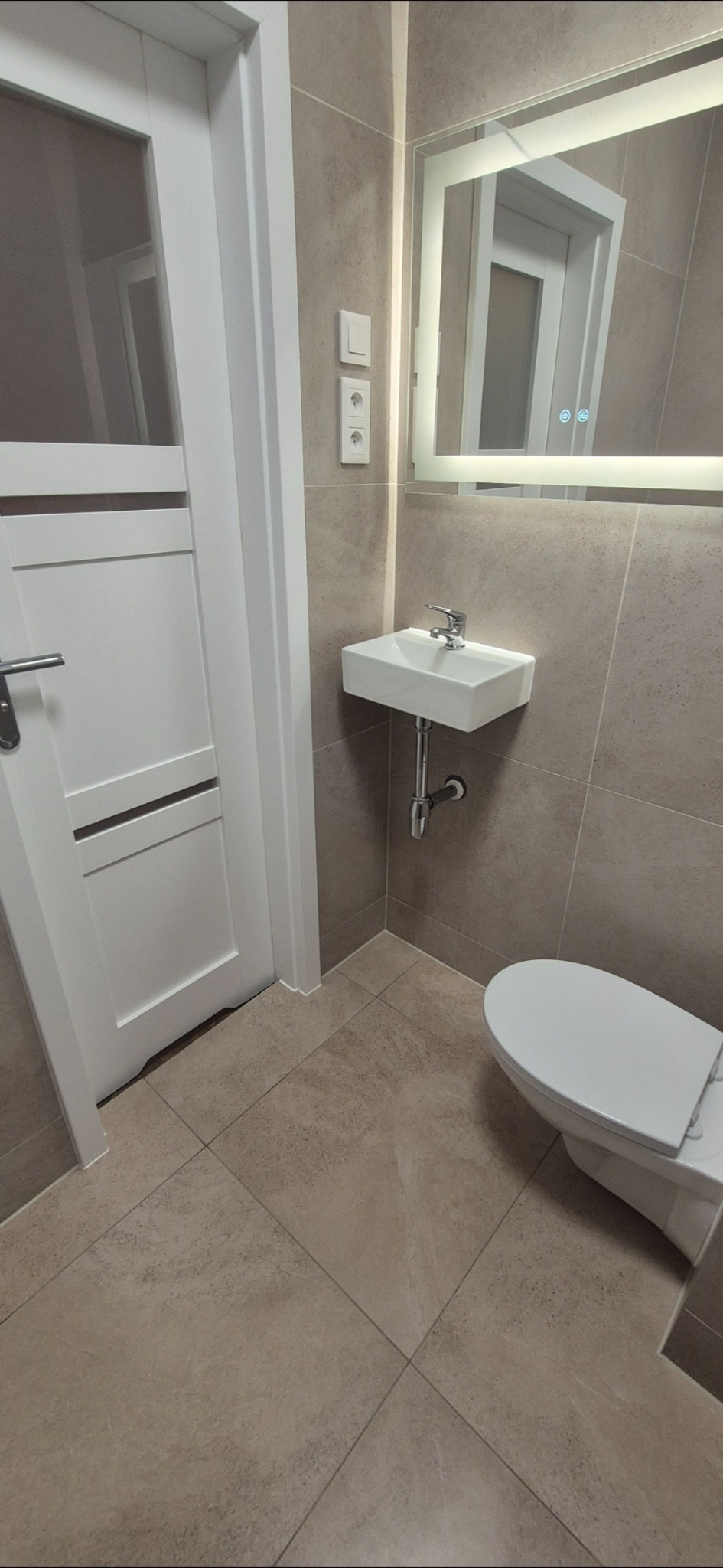 Nowoczesna łazienka w beżowych płytkach z podświetlanym lustrem, białą umywalką i toaletą. Widoczna biała, częściowo otwarta, lakierowana stolarka drzwiowa.