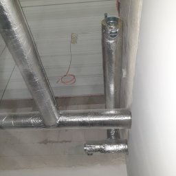 AIRMAX.SERVIS SPÓŁKA Z OGRANICZONĄ ODPOWIEDZIALNOŚCIĄ - Izolowane rury wentylacyjne pod sufitem z blachy trapezowej. Widoczne przewody elektryczne i fragment ściany z niedokończonym tynkiem.