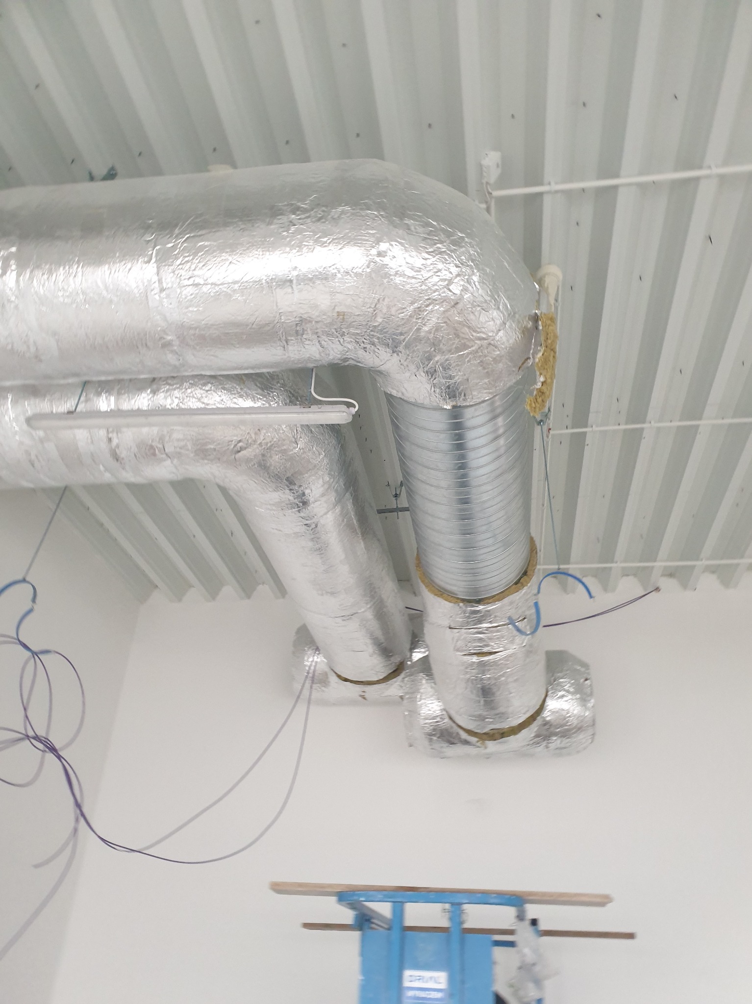 Srebrne, izolowane przewody wentylacyjne pod stropem z blachy trapezowej. Widoczne elementy montażowe i fragment podnośnika. Ujęcie z dołu.