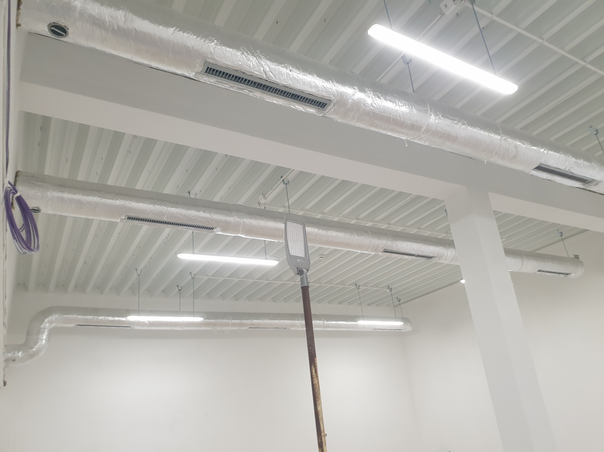 Białe wnętrze z widoczną instalacją wentylacyjną pod sufitem, rury owinięte folią aluminiową, kratki wentylacyjne i lampy sufitowe. Widok z dołu.