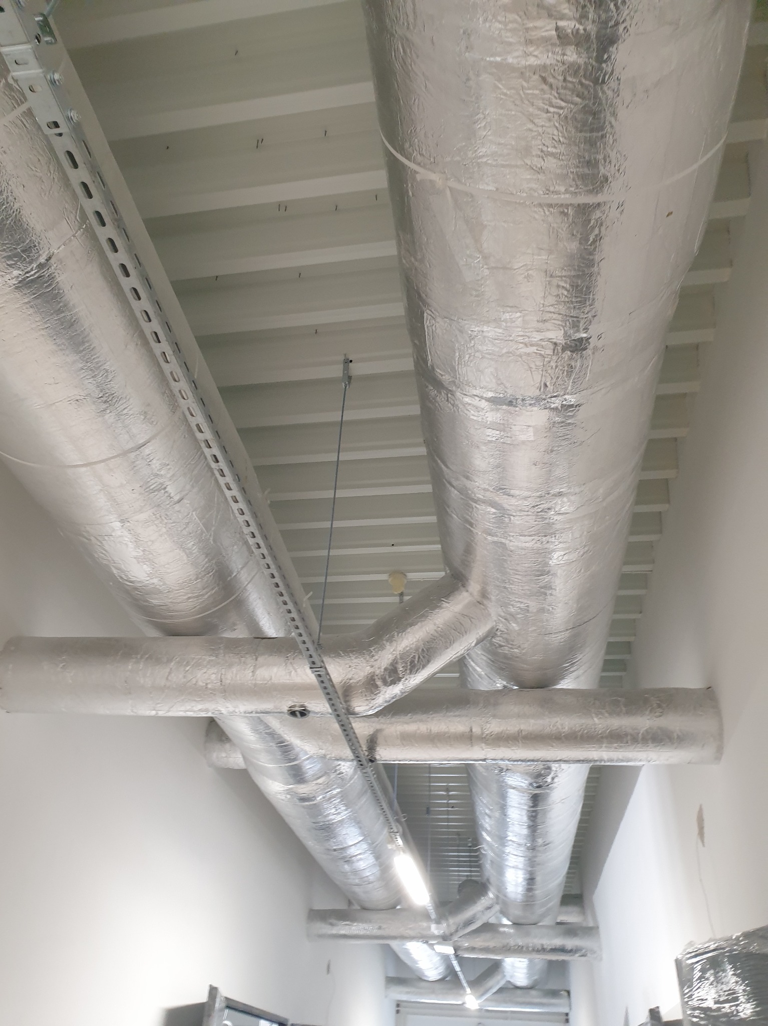 Srebrne, izolowane rury wentylacyjne pod sufitem, mocowane na metalowych szynach. Widok od dołu ukazuje rozgałęzienia i elementy instalacji w jasnym pomieszczeniu.