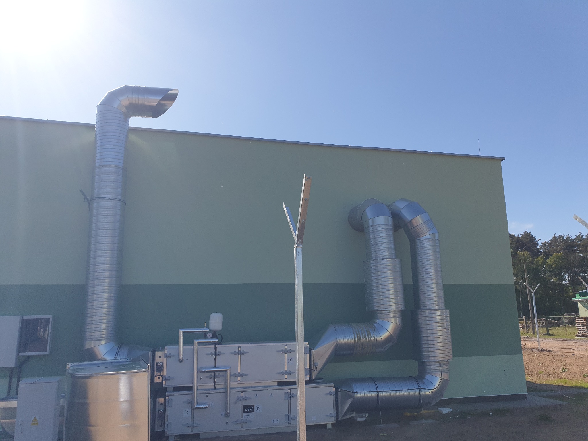 Zewnętrzny montaż instalacji wentylacyjnej z rurami na ścianie budynku o dwukolorowej elewacji w słoneczny dzień. Widoczne elementy systemu wentylacyjnego.