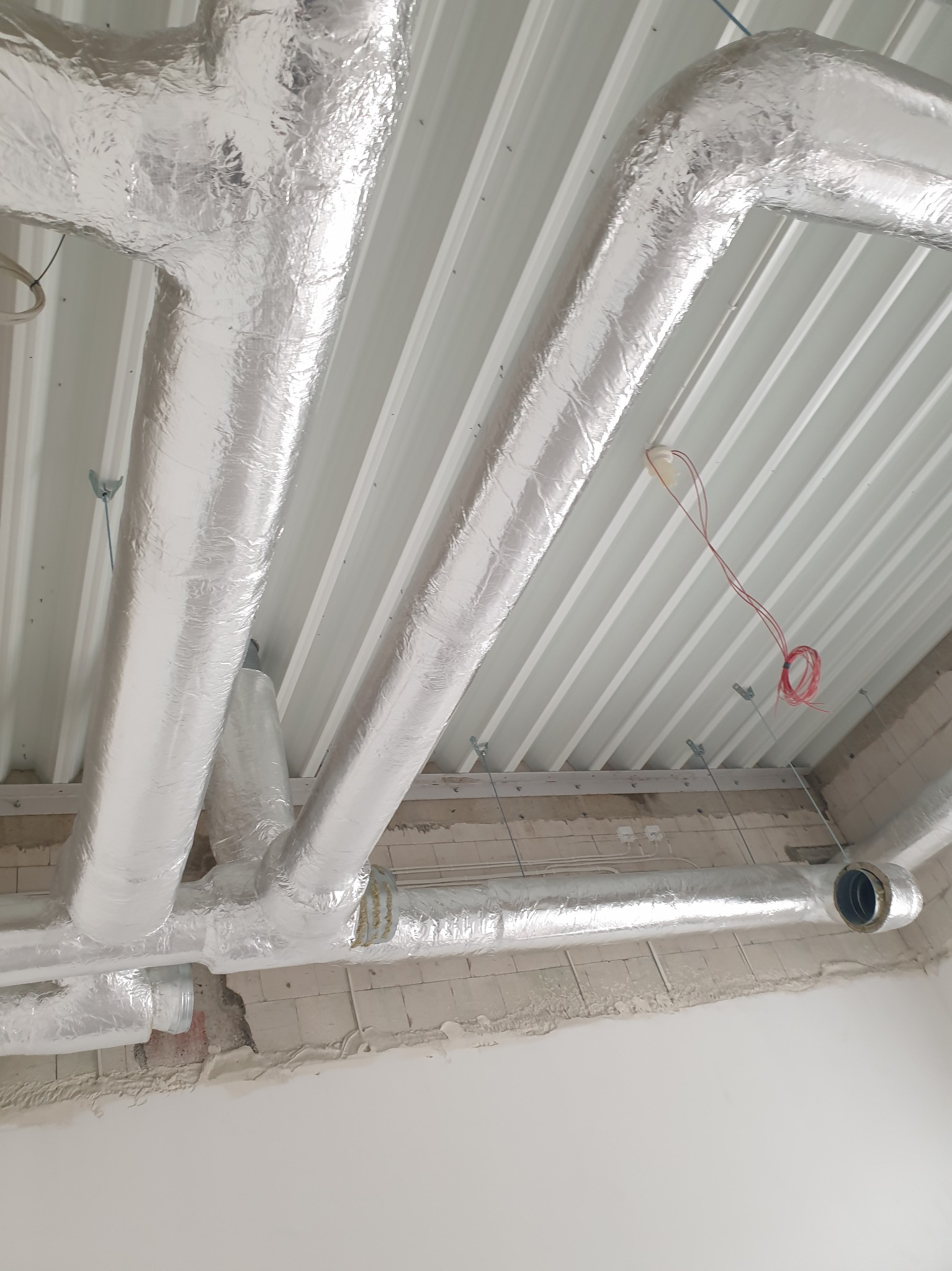 Srebrne, izolowane rury wentylacyjne zamontowane pod białym, żebrowanym sufitem, widoczne elementy instalacji elektrycznej i surowy stan wykończenia ścian.