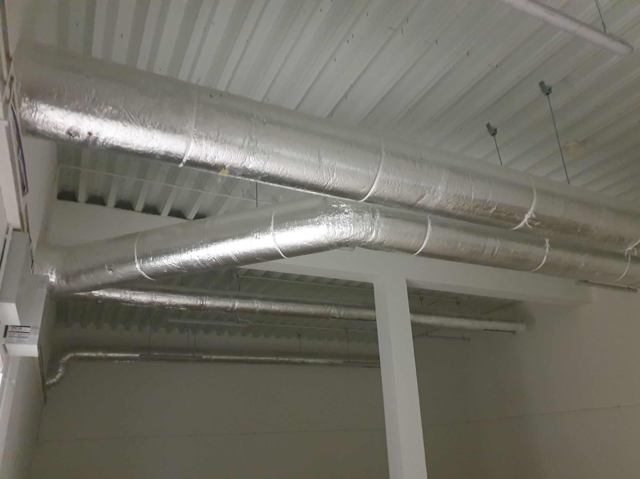 Szklane rury wentylacyjne z izolacją aluminiową zamontowane pod sufitem w jasnym pomieszczeniu. Widoczne elementy mocujące i fragmenty instalacji.